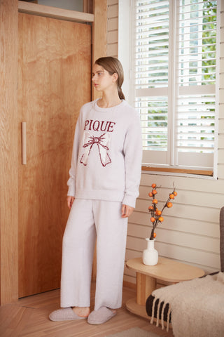 Woman wearing Ribbon Pullover Sweater & Long Pants Set by Gelato Pique, standing in a cozy room with slippers and plant decor.