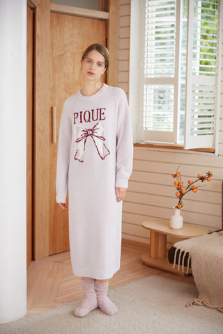 Woman wearing a long burgundy ribbon patterned jacquard dress in a cozy room setting with wooden accents.