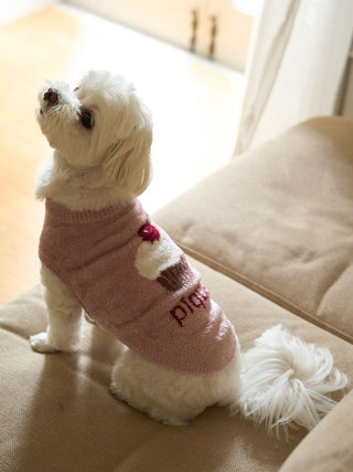 Small white dog wearing a pink CAT&DOG Cupcake sleeveless pet sweater, sitting on a beige sofa.