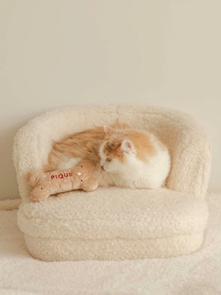 Cat lounging on a plush chair with a bone-shaped toy labeled 'PIQUE', in a cozy, neutral-toned setting.