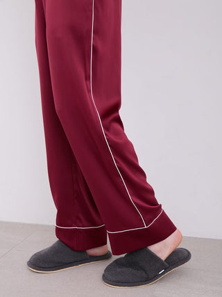 Men's burgundy piping satin pajama pants, silky texture with elegant white piping, worn with grey slippers, resting on a light floor.