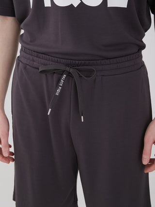 Unisex big logo lounge shorts with drawstring and logo waist strap, featuring knee-length relaxed fit and back pocket in dark fabric.