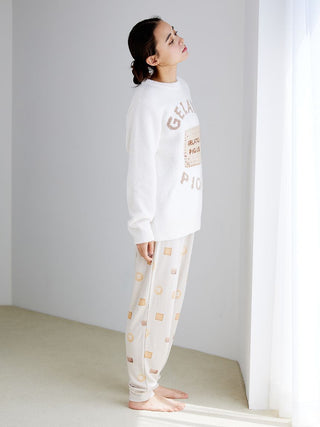Woman wearing biscuit pattern waffle pajama pants and sweater, standing near window with soft light.
