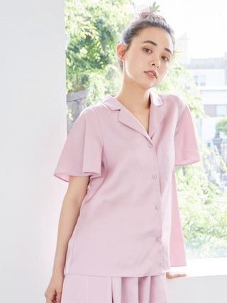 Woman wearing a light pink satin sleep shirt with button-up design, standing by a window with green foliage outside.