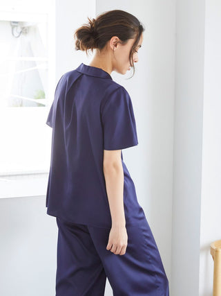 Woman wearing a navy satin sleep shirt and pants, standing by a window in a minimalist room.