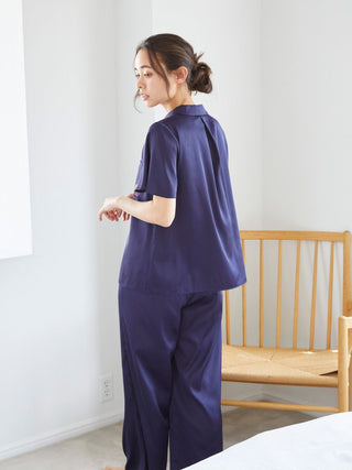Woman wearing a navy satin sleep shirt and pants, standing beside a wooden chair in a bright room.