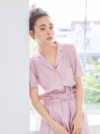 Woman wearing a strawberry embroidered satin sleep shirt, featuring a button-up design, standing by a window with greenery outside.