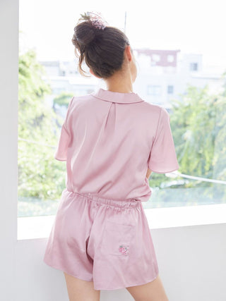 Woman wearing pink satin sleep shirt and shorts with strawberry embroidery, standing by a window.