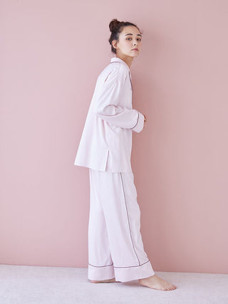 Woman wearing pink piping satin long sleeve pajama shirt and pants against a pink background.