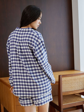 Woman wearing Gelato Pique checkered long sleeve sleep shirt and shorts, standing by a wooden chair indoors.