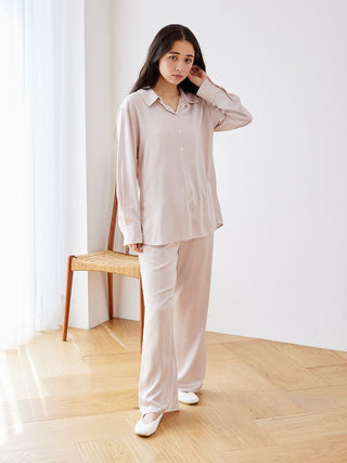 Woman wearing long sleeve satin sleepwear blouse and pants, standing in a bright room with a wooden chair and soft light.