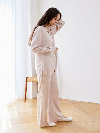 Woman wearing long sleeve satin sleepwear blouse and pants, standing in a bright room with wooden flooring, showcasing elegant design.