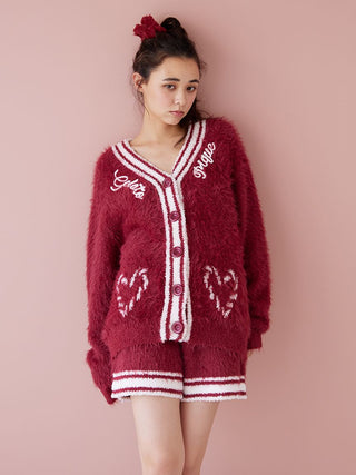 Woman in a cozy maroon cardigan and shorts set with heart patterns, wearing a matching fur hair clip, against a soft pink background.