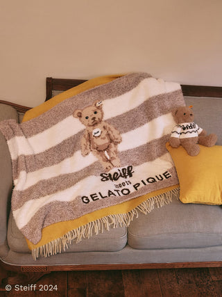 Steiff throw blanket with teddy bear design on a couch, beige and white striped with 'Gelato Pique' text