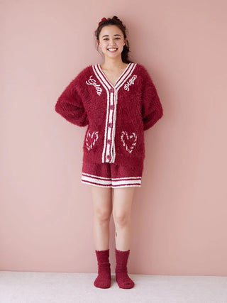 Woman wearing a cozy red fuzzy cardigan and shorts with matching socks, standing against a pink background, smiling.