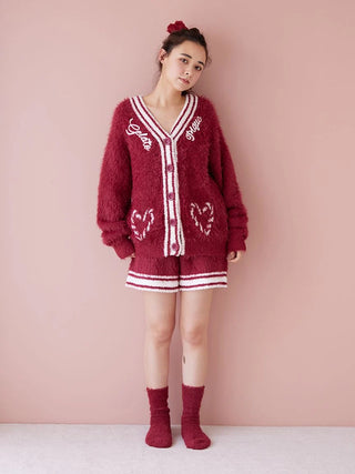 Woman wearing red cozy knit cardigan and shorts set, standing against pink background.