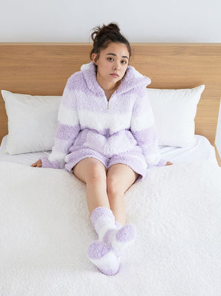 Woman wearing fluffy purple and white striped lounge shorts and hoodie, sitting on white bed with wooden headboard.