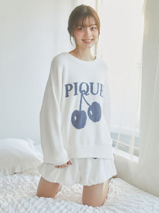 Woman wearing a cozy white sweater with cherry print, kneeling on bed, smiling, in bright, airy room.