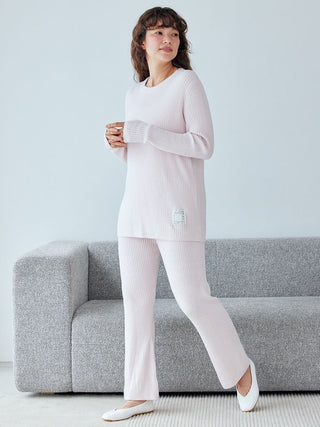 Woman wearing a pink ribbed pullover sweater and pants set while standing by a gray sofa.