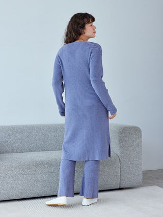 Back view of a woman wearing a blue ribbed long cardigan and matching pants, standing in a modern living room.