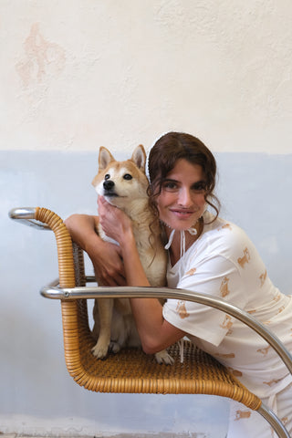 Woman in white Shiba Motif Pajama Pants by Gelato Pique USA, hugging a Shiba Inu dog on a wicker chair. Premium loungewear and sleepwear.