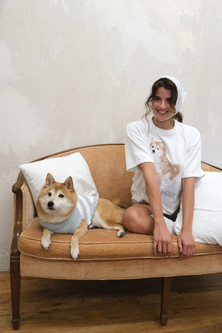 Young woman in white Shiba Inu-themed loungewear sitting on a sofa with a Shiba Inu dog wearing a blue sweater, Gelato Pique USA collection.