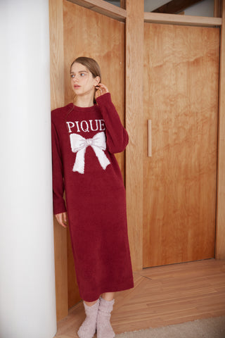 Woman wearing a burgundy Ribbon Jacquard Dress with white bow motif, standing by a wooden door. Cozy loungewear style.