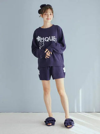 Woman wearing Airy Moco Flower Logo Jacquard Pullover Sweater and shorts in a cozy lounge setting.