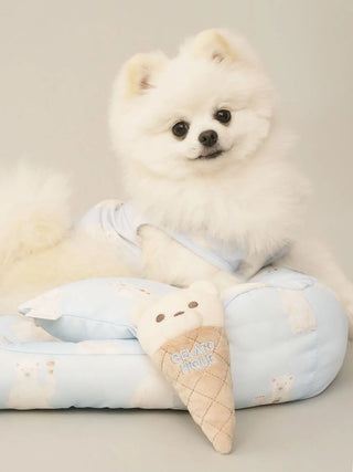 Fluffy white dog wearing light blue premium loungewear beside a Gelato Pique USA bear ice cream toy on a matching cushion.