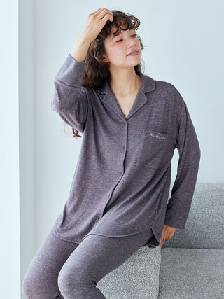 Woman wearing a cozy gray long sleeve pajama shirt and pants set, sitting on a couch, smiling, and touching her hair.