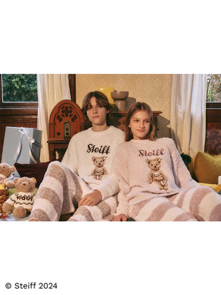 Couple wearing Steiff Gelato 2 Border Long Pants with teddy bear design, seated in a cozy room with plush fabric and striped patterns.