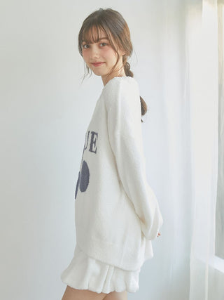 Model wearing a cozy white sweater with graphic print, showcasing a relaxed style in a softly lit room.