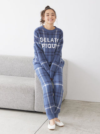 Woman wearing a blue checkered pajama set, sitting on a gray sofa. Cozy loungewear with Gelato Pique logo in a relaxed fit design.