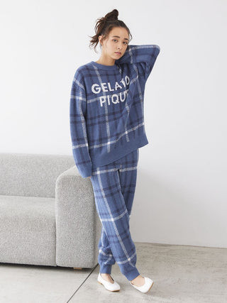 Woman wearing Gelato Pique checkered pajama set in blue with relaxed fit, standing next to a gray couch, showcasing cozy loungewear.