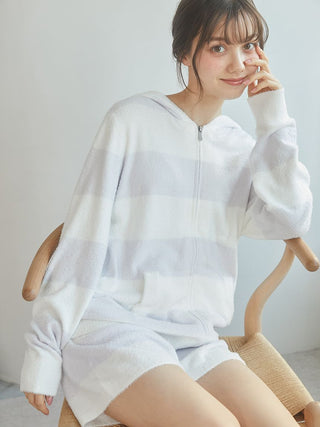 Young woman wearing a light striped zip-up hoodie and shorts, sitting on a chair with a cheerful expression.