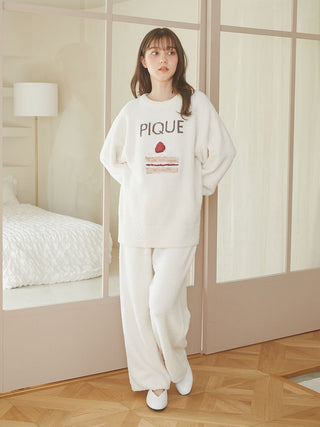 Woman wearing Cake Jacquard Pullover Sweater with cake motif and 'PIQUE' lettering, standing in cozy loungewear beside a bedroom.