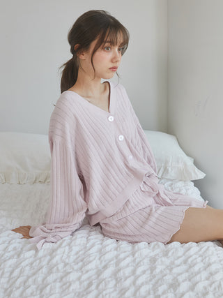 Woman wearing pink ribbed knit shorts and cardigan sits on a bed.